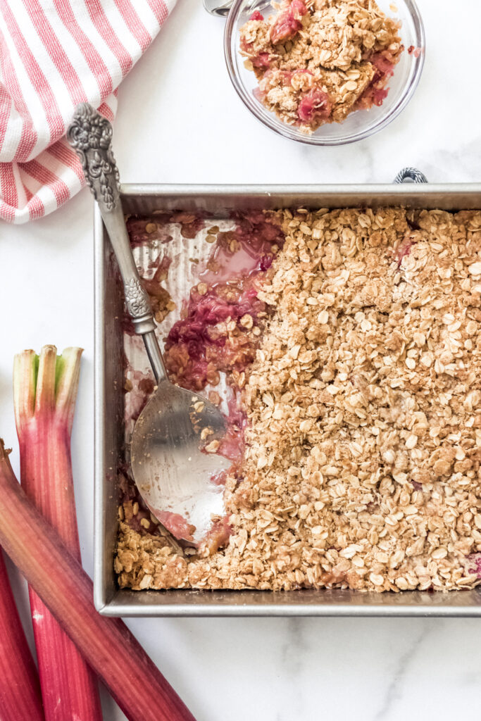 Rhubarb crisp in a baking dish with a serving spoon next to stalks of fresh rhubarb.