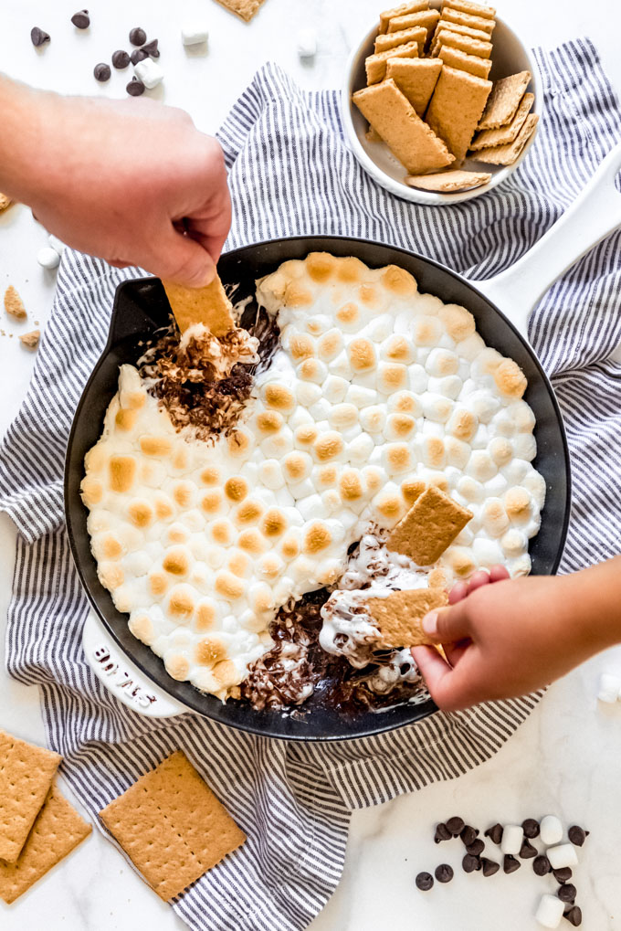 Two hands holding graham crackers to scoop melted s'mores dip from a skillet.