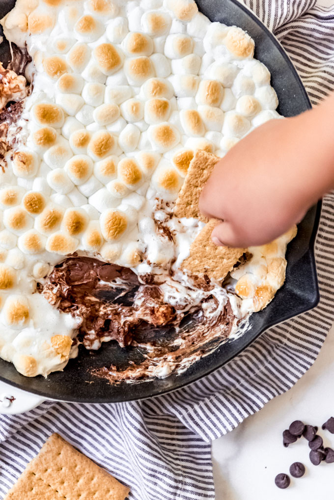 A hand using a graham cracker to scoop s'mores dip from a skillet.