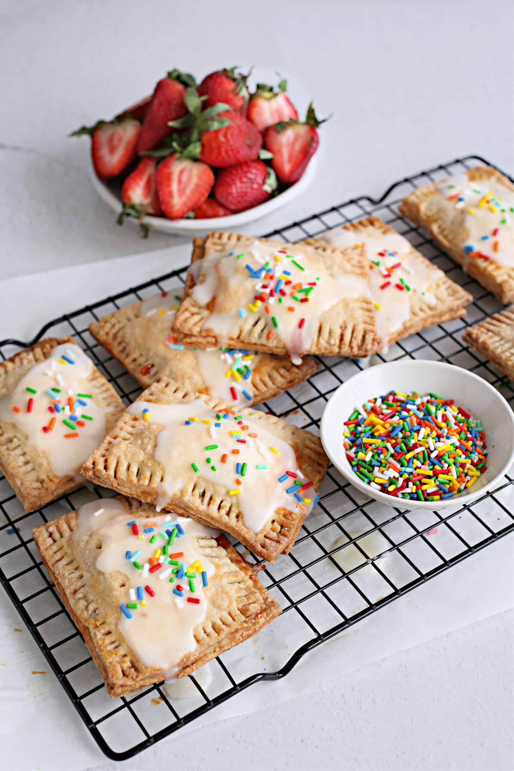 These pastries feature a golden, flaky crust filled with sweet fruit jam, baked to perfection on a wire rack and topped with glaze and confetti sprinkles. A bowl of sprinkles and a bowl of strawberries sit nearby.