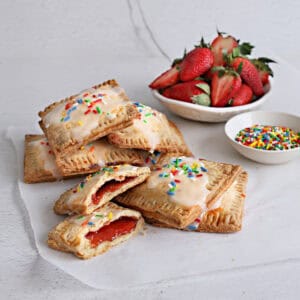 These pastries feature a golden, flaky crust filled with sweet fruit jam, baked to perfection on a piece of parchment paper and topped with glaze and confetti sprinkles. A bowl of sprinkles and a bowl of strawberries sit nearby.