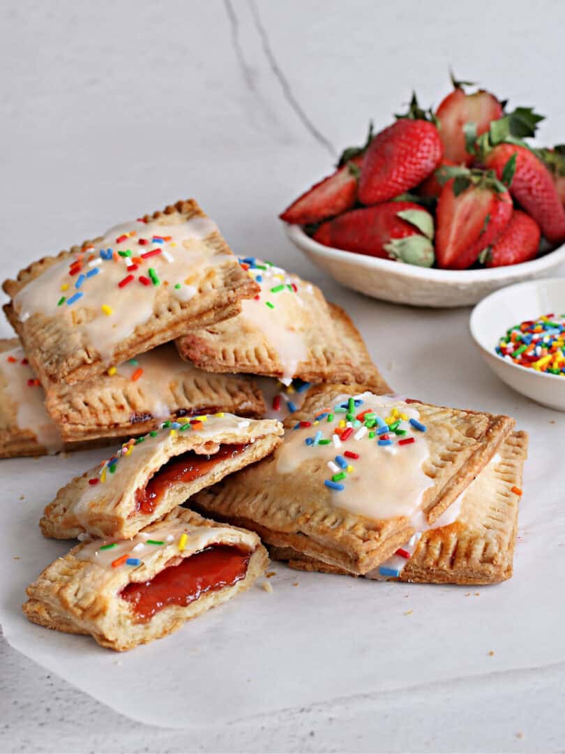 These pastries feature a golden, flaky crust filled with sweet fruit jam, baked to perfection on a piece of parchment paper and topped with glaze and confetti sprinkles. A bowl of sprinkles and a bowl of strawberries sit nearby.