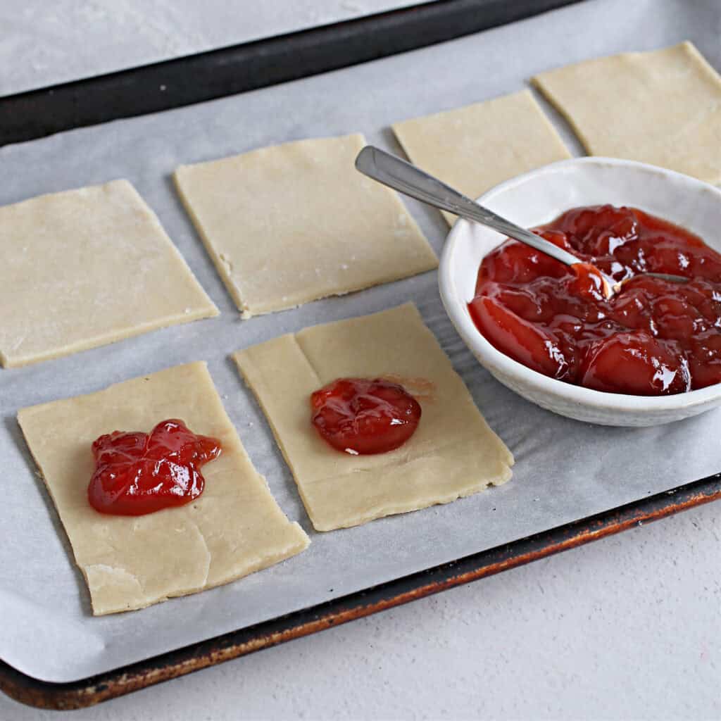 Image shows dough for strawberry pop tarts, a rolling pin, and strawberry jam in a bowl, ready for baking on a white countertop.
