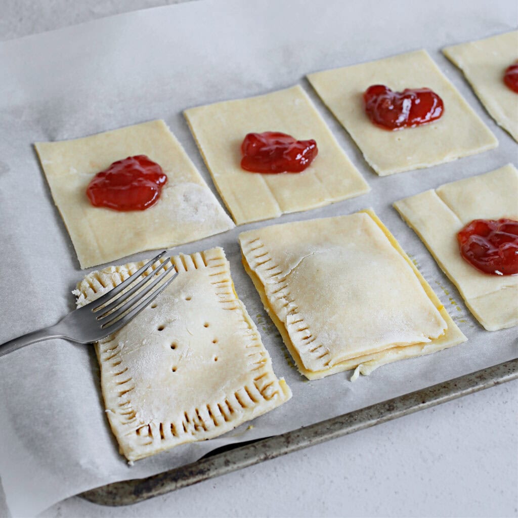 Pastry squares with strawberry jam filling ready for baking, on parchment paper.